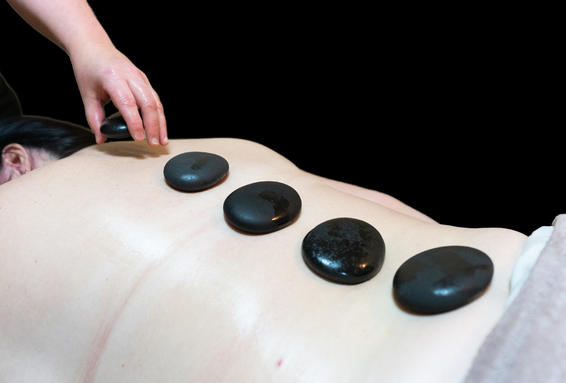 Cedar Meadows Spa & Resort Person receiving a hot stone massage with smooth, black stones placed along their back, against a black background. - Timmins, Ontario