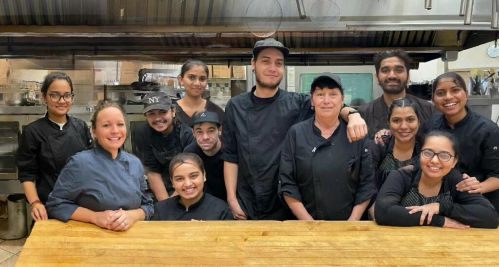 Cedar Meadows Spa & Resort A diverse group of kitchen staff in black uniforms pose together behind a wooden counter in a commercial kitchen. - Timmins, Ontario