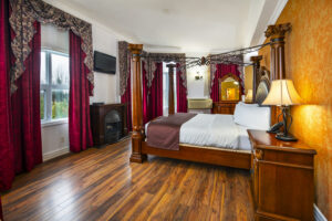 Cedar Meadows Spa & Resort A bedroom with a four-poster bed, wooden furniture, a fireplace, a TV, and red curtains. The lamps on the nightstands are lit. - Timmins, Ontario