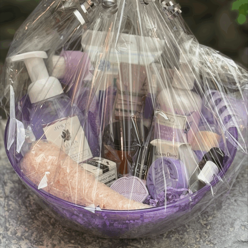 Cedar Meadows Spa & Resort A gift basket wrapped in clear plastic, containing various bath and body products in purple and white packaging, set on a gray surface. - Timmins, Ontario