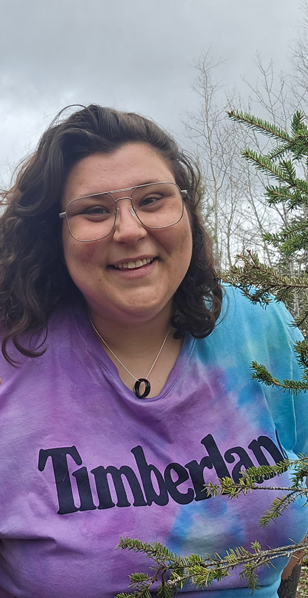 Cedar Meadows Spa & Resort A person with long brown hair and glasses smiles outdoors, wearing a tie-dye Timberland shirt and a necklace, with pine branches and cloudy sky in the background. - Timmins, Ontario