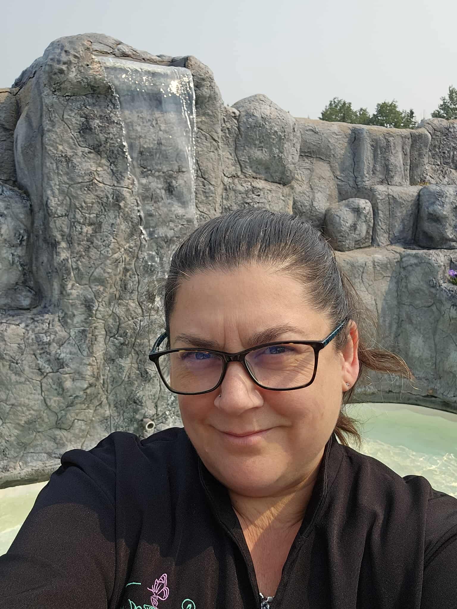 Cedar Meadows Spa & Resort A person wearing glasses and a black jacket takes a selfie in front of an artificial rock waterfall at a pool or water park. - Timmins, Ontario