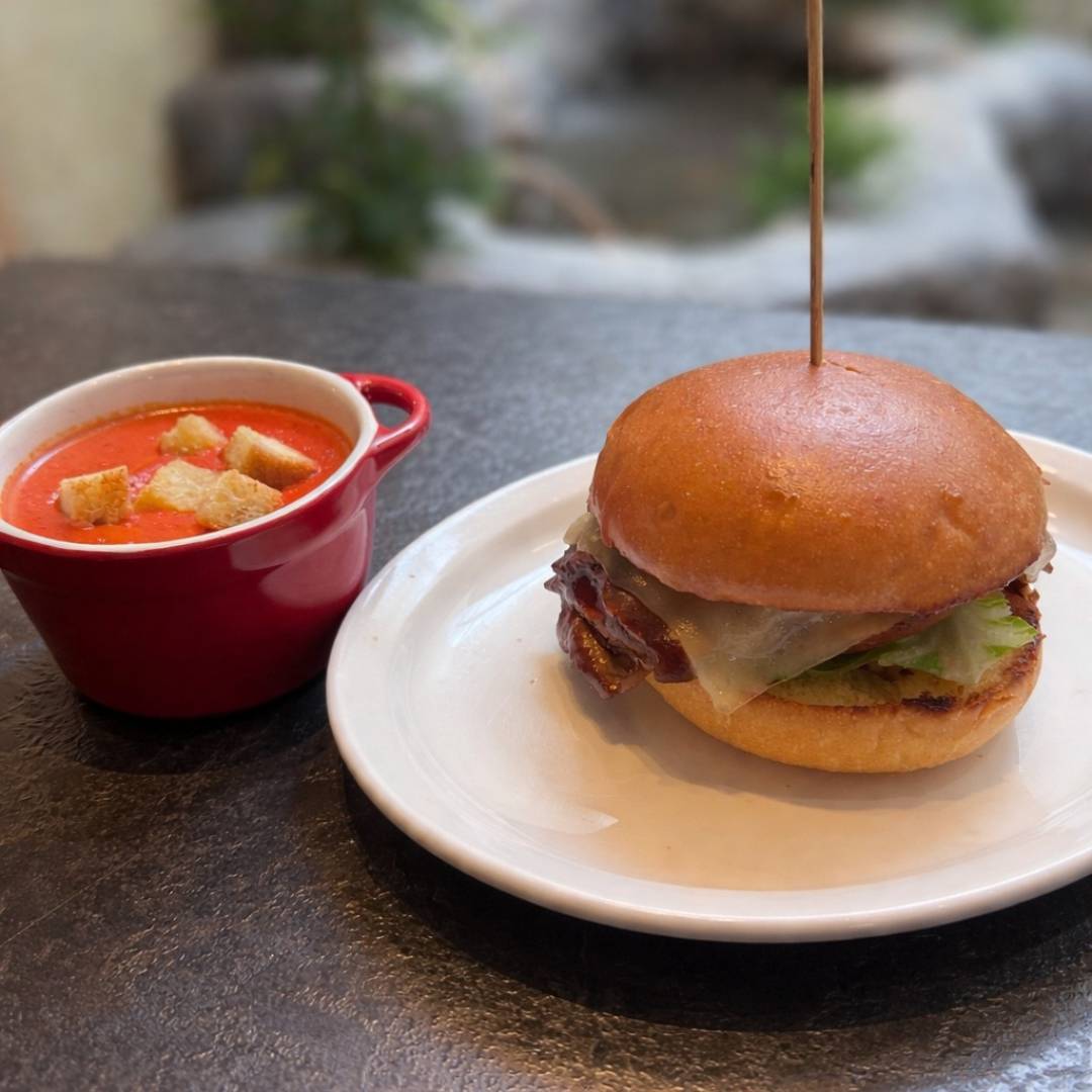 Cedar Meadows Spa & Resort A cheeseburger with lettuce and bacon on a white plate next to a red cup of tomato soup topped with croutons. - Timmins, Ontario
