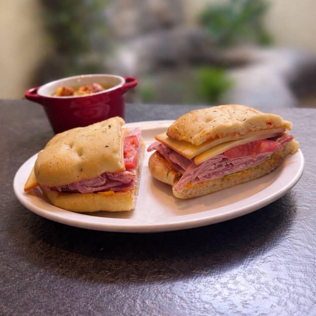 Cedar Meadows Spa & Resort A sandwich with ham, cheese, and tomato on focaccia bread, cut in half and served on a white plate, with a small dish of food in the background. - Timmins, Ontario