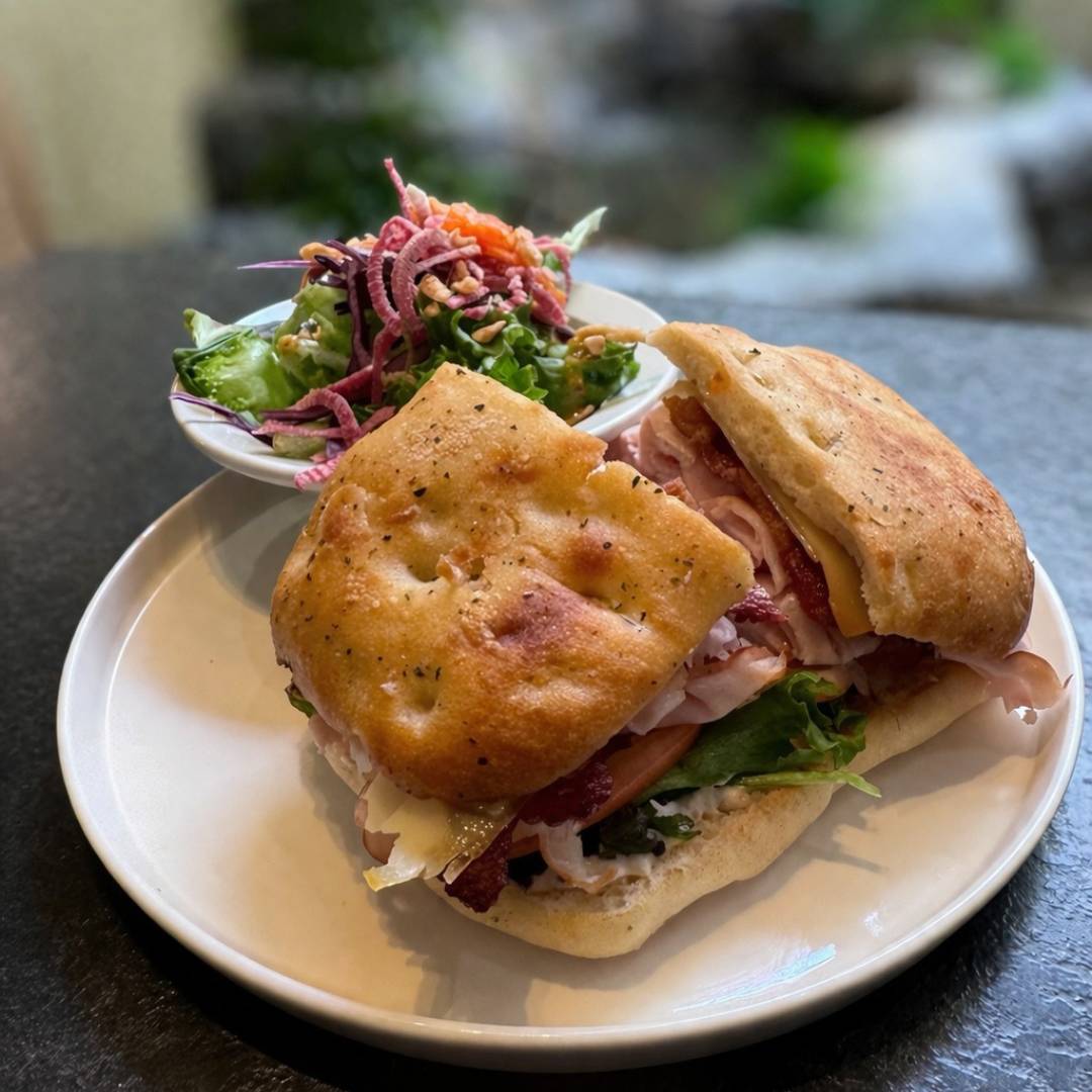 Cedar Meadows Spa & Resort A focaccia sandwich filled with lettuce, ham, cheese, and tomato is served on a white plate with a side of mixed salad topped with shredded vegetables. - Timmins, Ontario