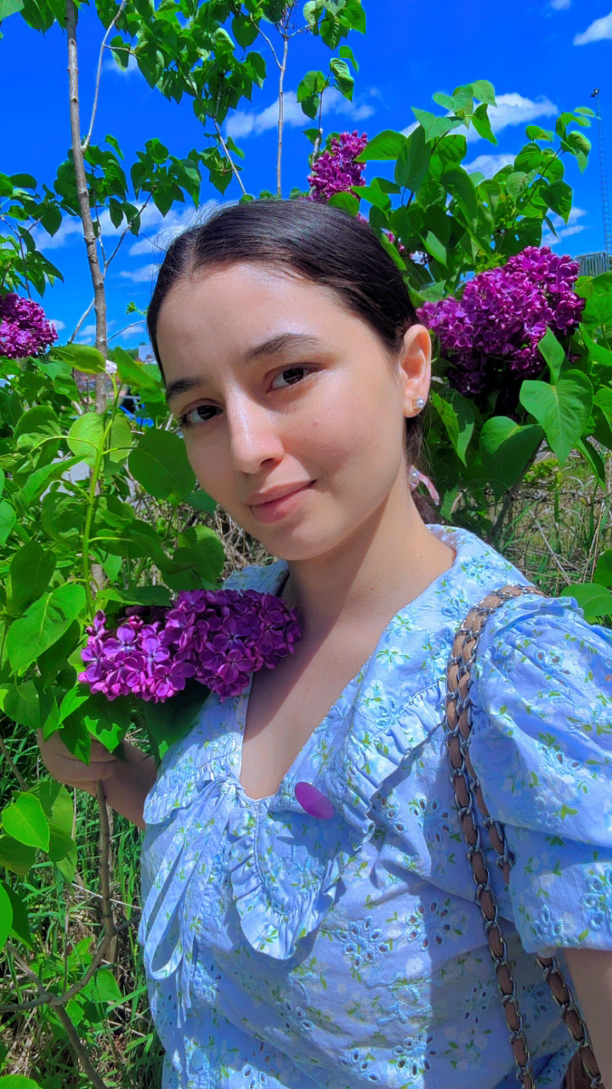 Cedar Meadows Spa & Resort A woman in a light blue floral dress stands among green leaves and purple flowers, looking at the camera on a sunny day. - Timmins, Ontario